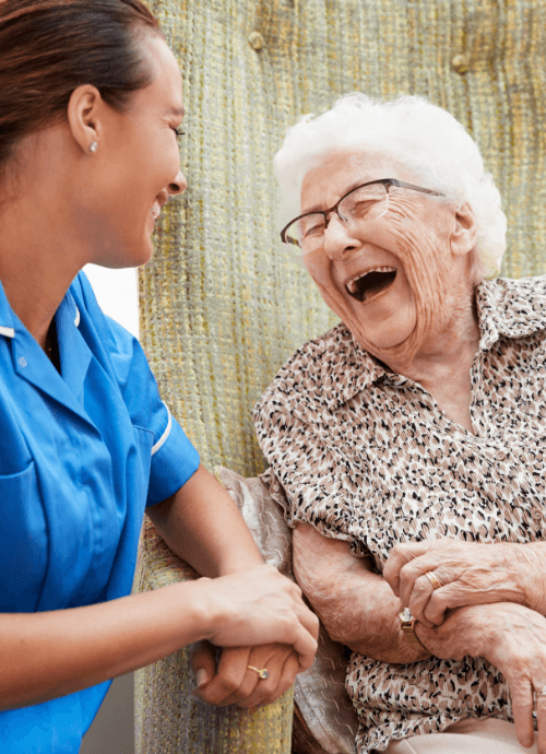Femme âgée assise dans un fauteuil et riant avec une infirmière dans une maison de retraite.